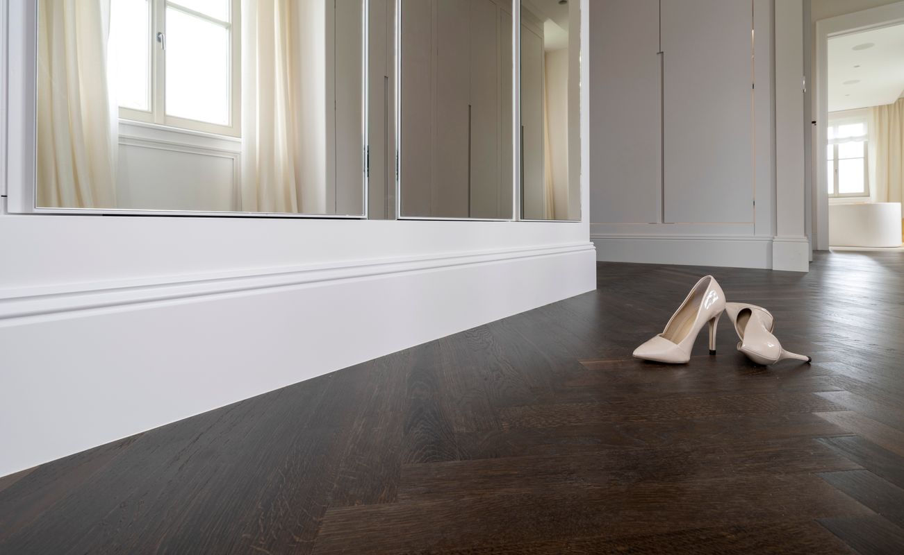 Beige High Heels stehen auf einem dunklen Holzboden vor einem großen Spiegel in einem hellen Raum mit einem Fenster.