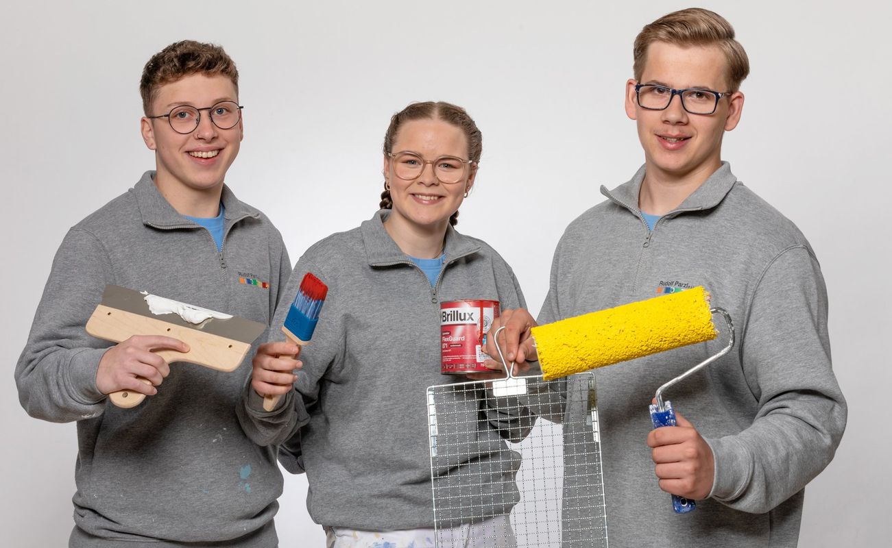 Drei Personen in grauen Sweatshirts halten Malerwerkzeuge: Spachtel, Farbroller und Farbdose. Hintergrund ist neutral.