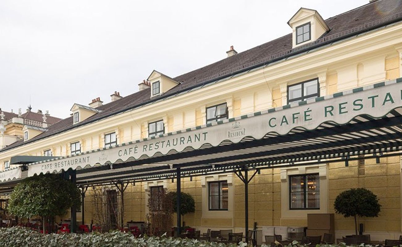 Ein elegantes Café-Restaurant mit gelber Fassade und Markisen, umgeben von Bäumen und Pflanzen.
